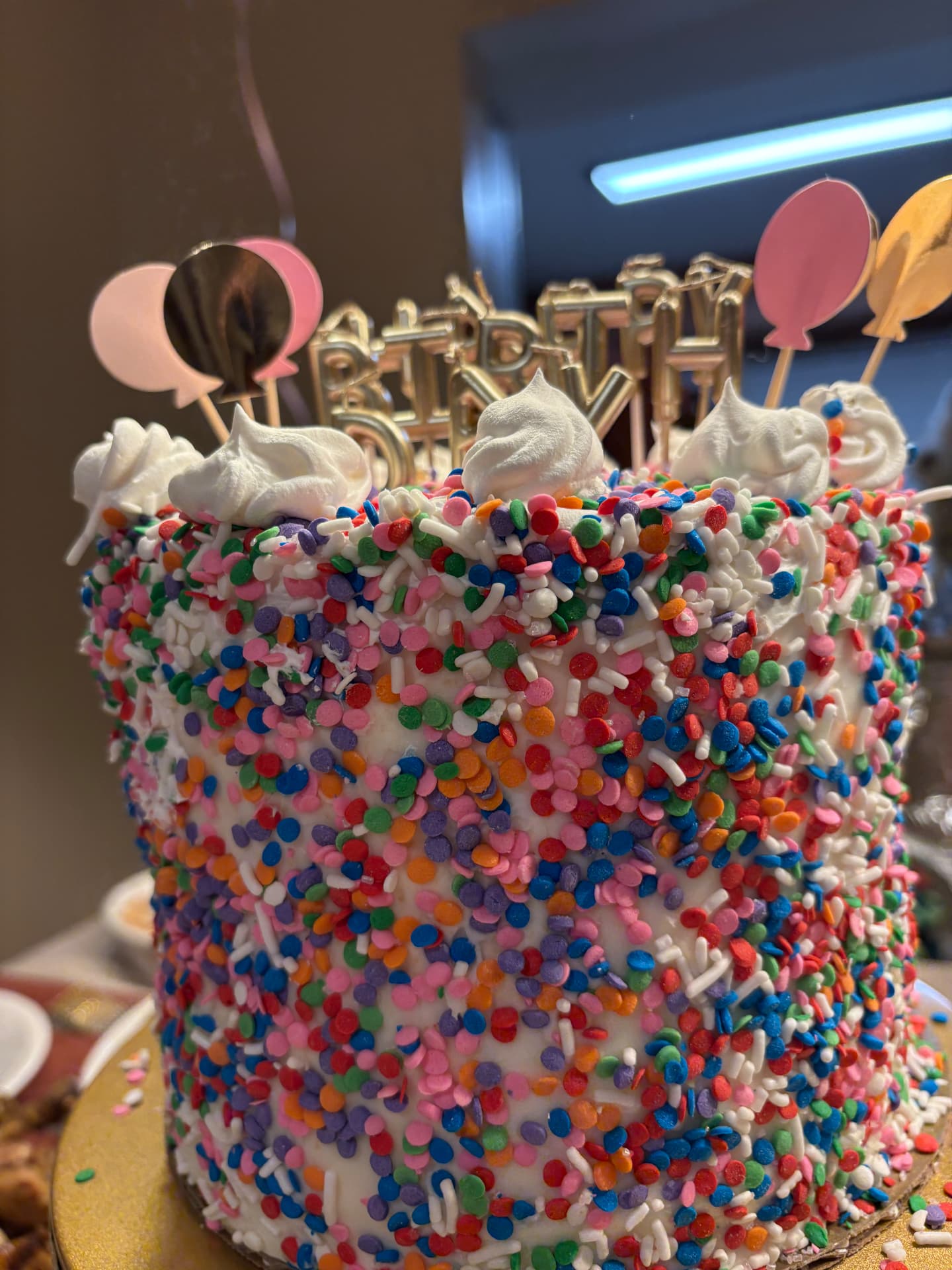 Tall birthday cake covered in rainbow sprinkles with whipped frosting swirls and balloon toppers